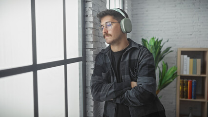 A pensive young hispanic man with a moustache and headphones stands indoors, arms crossed, against...