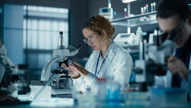 Cinematic Footage of a Female Bioengineer Looking at DNA Samples Under a Microscope in a Modern Applied Science Laboratory. Portrait of a Young Lab Engineer in White Coat Inventing New Medical Drugs