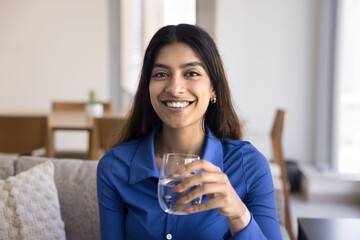 Positive beautiful healthy young Indian woman drinking clear water, holding glass with cold beverage, caring for health, nutrition, aqua balance, hydration, looking at camera with toothy smile - 785297377