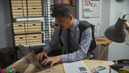 Hispanic man working on a laptop at a cluttered detective office with evidence scattered around.