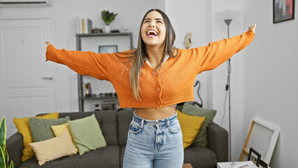 Joyful hispanic woman laughing in cozy apartment living room with vibrant orange sweater