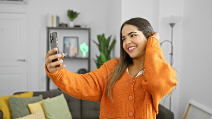 A cheerful young hispanic woman takes a selfie at home, exuding beauty and positivity in a cozy...