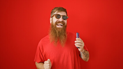 Cheerful young irish male with beard, dancing over isolated red wall, joyfully listening to funny tunes on mobile phone. handsome redhead guy smiling, holding cellphone enjoying online music.