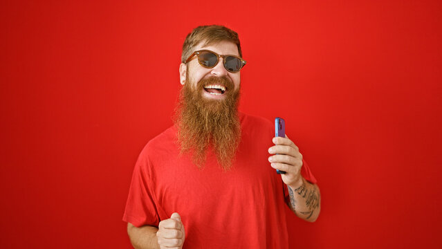 Cheerful young irish male with beard, dancing over isolated red wall, joyfully listening to funny tunes on mobile phone. handsome redhead guy smiling, holding cellphone enjoying online music.