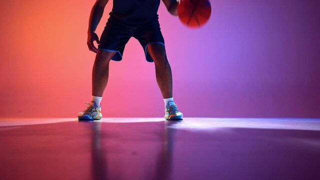 Dynamic display of athleticism. Skillful male basketball player showing dribbling technique with ball on gradient red purple background in neon light. Concept of professional sport, competition, game