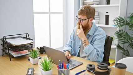 Confident young redhead man working joyfully on laptop at office, exuding confidence as business...