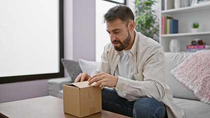 Handsome young hispanic man, engrossed and serious, unpacking delivery from cardboard box while sitting in living room sofa, portrait of chilling on a relaxed day at home. - obrazy, fototapety, plakaty
