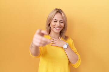 Young caucasian woman wearing yellow sweater laughing at you, pointing finger to the camera with hand over body, shame expression