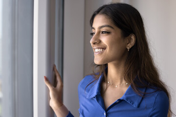 Happy dreamy beautiful 20s Indian girl with perfect toothy smile and stylish nose stud looking at window away, smiling, thinking, daydreaming, planning future, enjoying view