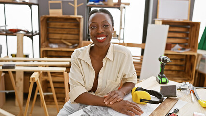 Smiling african american woman carpenter confidently sitting at the carpentry table in her busy,...