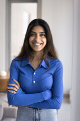 Cheerful attractive young Indian woman standing at home with hands folded, looking at camera, smiling, posing for head shot. Successful confident beautiful 20s girl vertical portrait