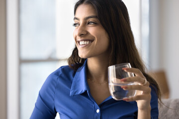 Positive beautiful 20s Indian girl holding glass of clear water, looking away, thinking on healthy lifestyle, diet, hydration balance, skincare, drinking cold beverage at home
