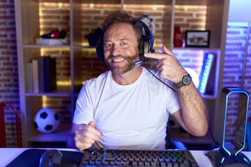 Middle age man with beard playing video games wearing headphones pointing with hand finger to face and nose, smiling cheerful. beauty concept