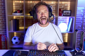 Middle age man with beard playing video games wearing headphones sticking tongue out happy with...
