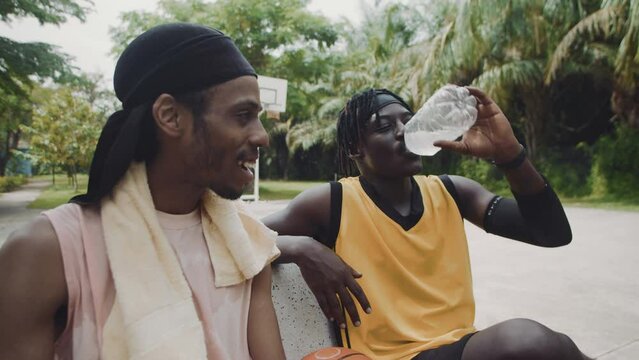Medium shot of two black male friends resting on bench, drinking water and chatting after basketball training outdoors