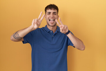 Young hispanic man standing over yellow background smiling with tongue out showing fingers of both...