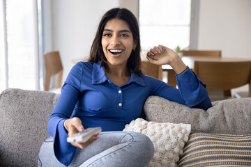 Excited positive young Indian woman watching TV show, movie, sport game, football match broadcasting, holding remote control device, sitting on home couch, smiling, shouting for joy - 785302578