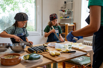 Twin sisters making maki and sushi dressed in a denim apron and cooking hat. The unrecognisable mother teaching and watching them. © Cristina