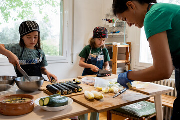 Twin sisters making maki and sushi dressed in a denim apron and cooking hat. The mother in front preparing endives and smiling.