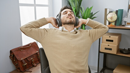 A relaxed young hispanic man with a beard enjoys music with headphones in a modern office setting.