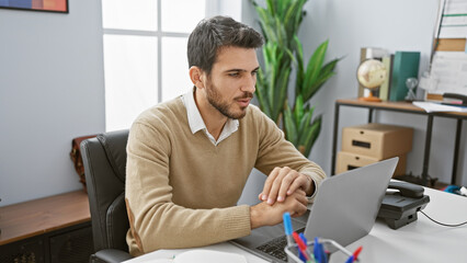 Handsome hispanic man with beard working on laptop in modern office environment.