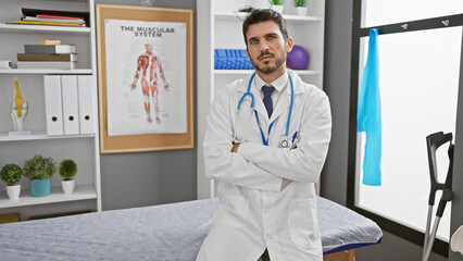 Handsome young man with beard, wearing lab coat, stands in clinic interior with arms crossed.