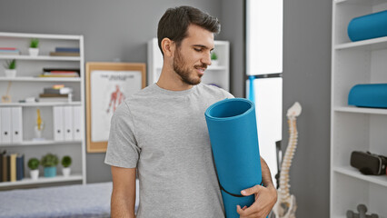 Handsome young hispanic man holding a yoga mat in a modern rehab clinic interior.