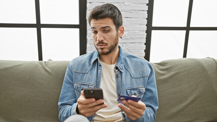 Handsome young adult hispanic man with beard using smartphone and holding credit card indoors at...