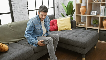 A young hispanic man with a beard in pain at home, holding his leg in a stylish interior living room