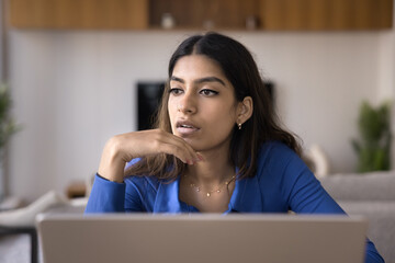 Thoughtful young Indian freelance entrepreneur woman working at laptop at home, looking away in deep thoughts, thinking on creative idea for project, business plan, strategy
