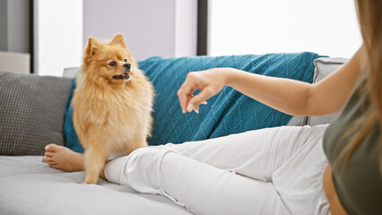 Heartwarming indoor portrait of a young hispanic woman playfully bonding with her lovable dog, lying on a sofa in her cozy living room at home, radiating happiness and touch of affection.