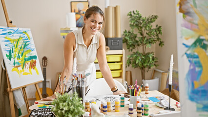 Confident young hispanic woman artist, laughing and enjoying her painting hobby in an art studio,...