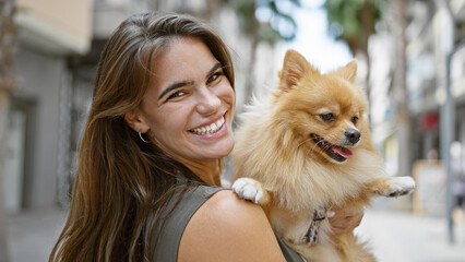 Cheerful young hispanic woman confidently standing on the city street, her beautiful smile radiating happiness as she hugs her adorable dog, their joy plainly visible under the sunny outdoors.