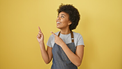 Cheerful african american woman with afro hair pointing upwards against a yellow background,...