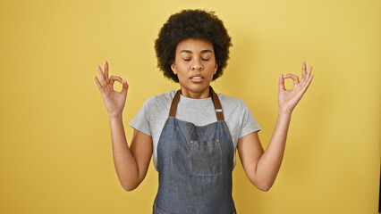 A serene adult black woman meditating in a denim apron against a yellow background.