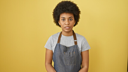 African american woman with curly hair wearing a denim apron stands against a yellow background,...