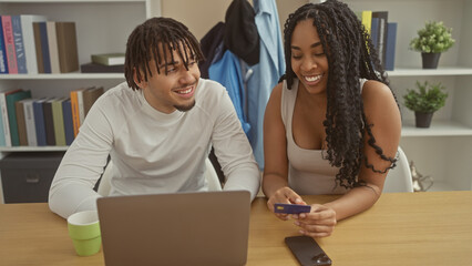 A smiling man and woman, casually dressed, are looking at a laptop with a credit card, in a modern living room.
