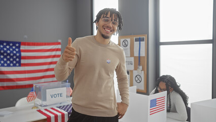 Smiling young man with 'i voted' sticker giving thumbs-up in front of american flag at a voting...