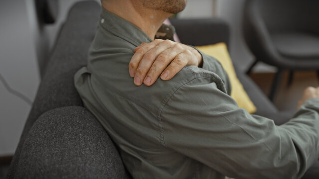 Young hispanic man with a beard grasping his shoulder in pain, seated indoors in a living room, portraying discomfort and aching.