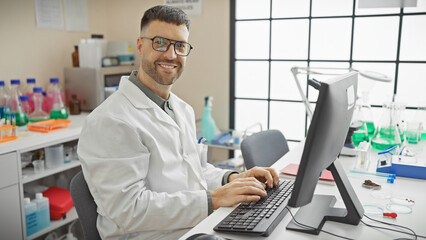 A smiling handsome hispanic man in a lab coat typing at a computer in a lab with scientific...