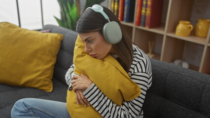 A pensive young woman wearing headphones cuddles a cushion on a sofa, exhibiting comfort and...