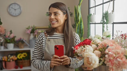 Attractive young hispanic woman with smartphone smiling in a floral decorated cozy interior room