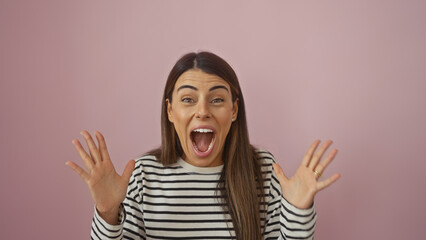 A cheerful young hispanic woman in a striped shirt expressing excitement against a pink isolated...