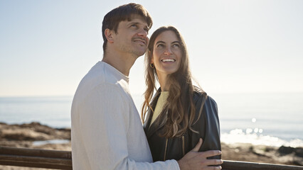 A smiling couple embraces on a sunny beachside promenade, gazing into the distance.