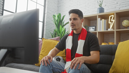 Hispanic man watching tv in a cozy living room, wearing a casual outfit and a sports scarf, with a...