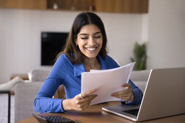 Cheerful satisfied Indian freelance business woman doing accounting job at home, reading paper financial reports, bills, tax invoices, counting profit, income from investment, smiling, laughing