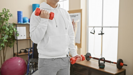 A man in a white hoodie exercising with dumbbells in a bright physiotherapy clinic interior.