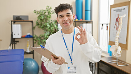 Handsome man with a clipboard giving an ok sign in a physiotherapy clinic