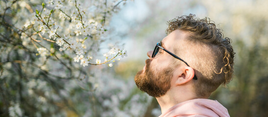 Banner portrait of curly millennial man inhales the fragrance of spring flowers of blooming jasmine...