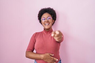 Beautiful african woman with curly hair standing over pink background laughing at you, pointing...
